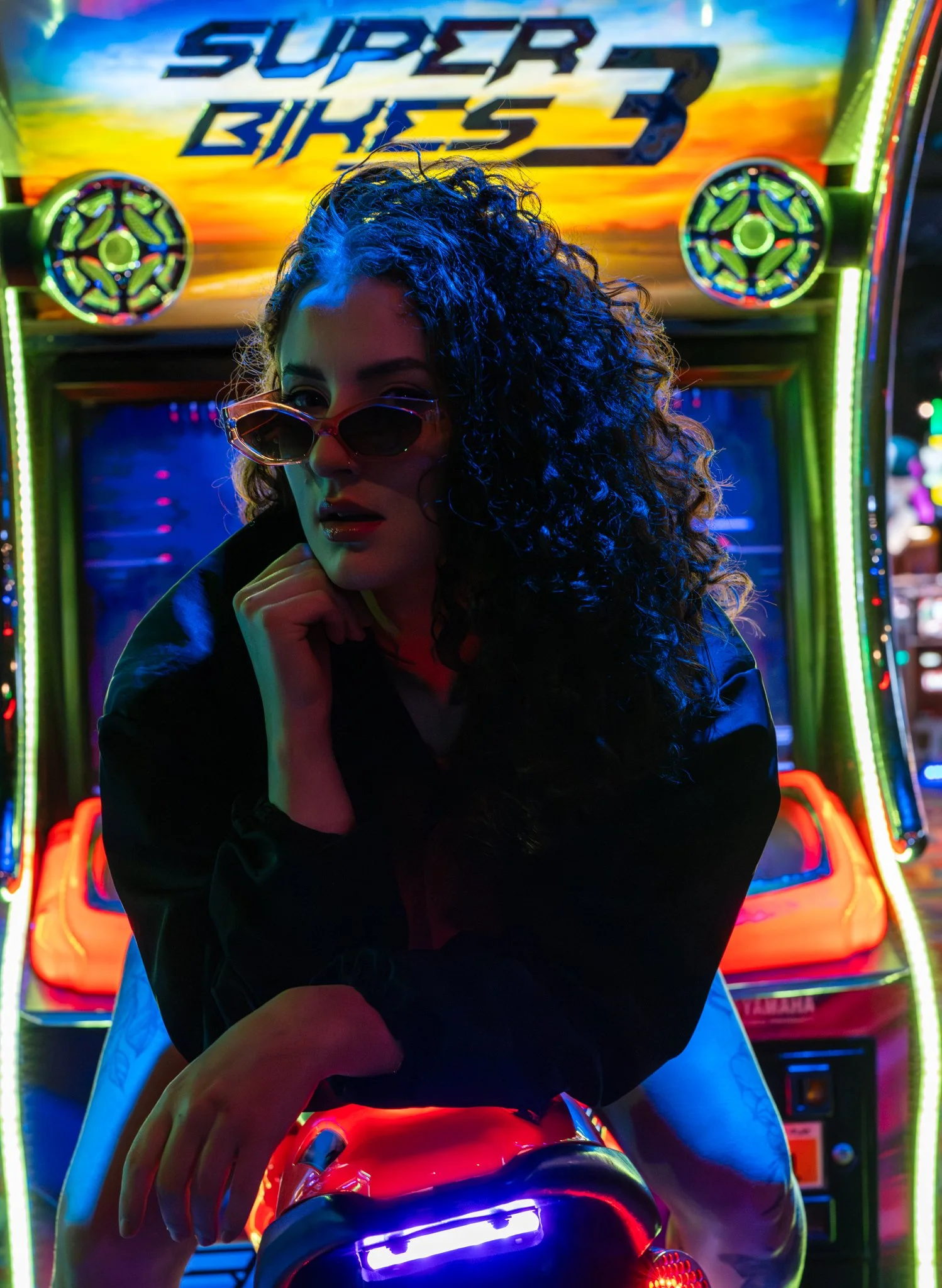 A woman with curly hair wearing sunglasses poses in front of a neon-lit arcade game called 'Super Bikes' at night. photographed in Walton, essex in the arcades on the pier. Anne Wills Photography 