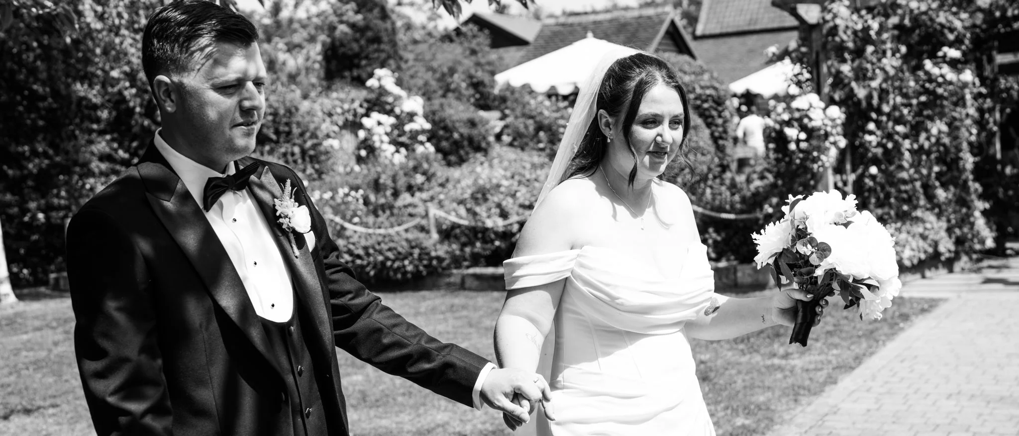 A black and white photo of a bride and groom holding hands outdoors during a wedding ceremony. The bride is holding a bouquet of flowers and wearing an off-the-shoulder wedding dress, with a veil. The groom is dressed in a tuxedo with a bow tie and a