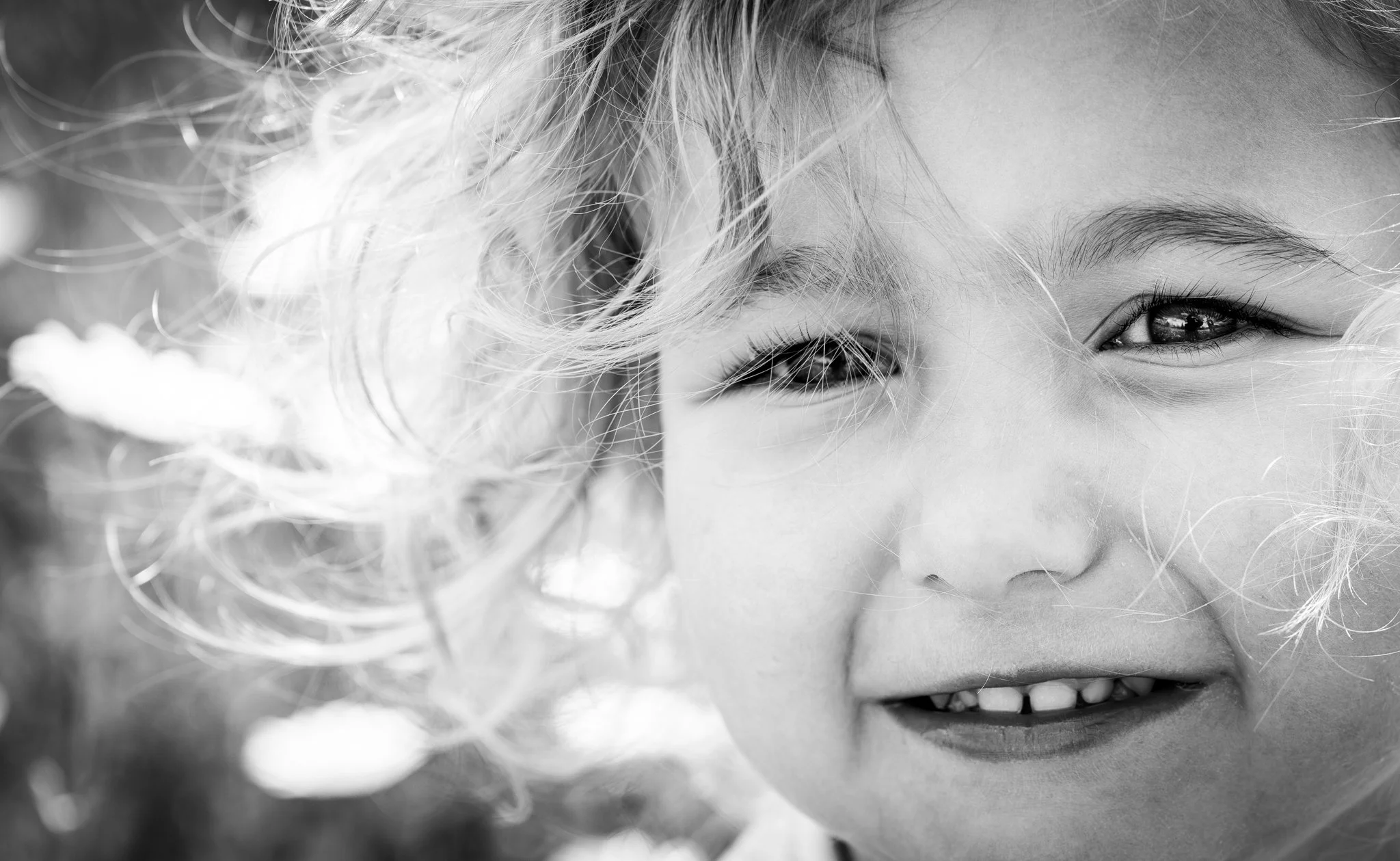 Close-up of a smiling girl with curly hair, showing her eyes and teeth, in black and white.  
Anne Wills Photography. Braintree, Essex