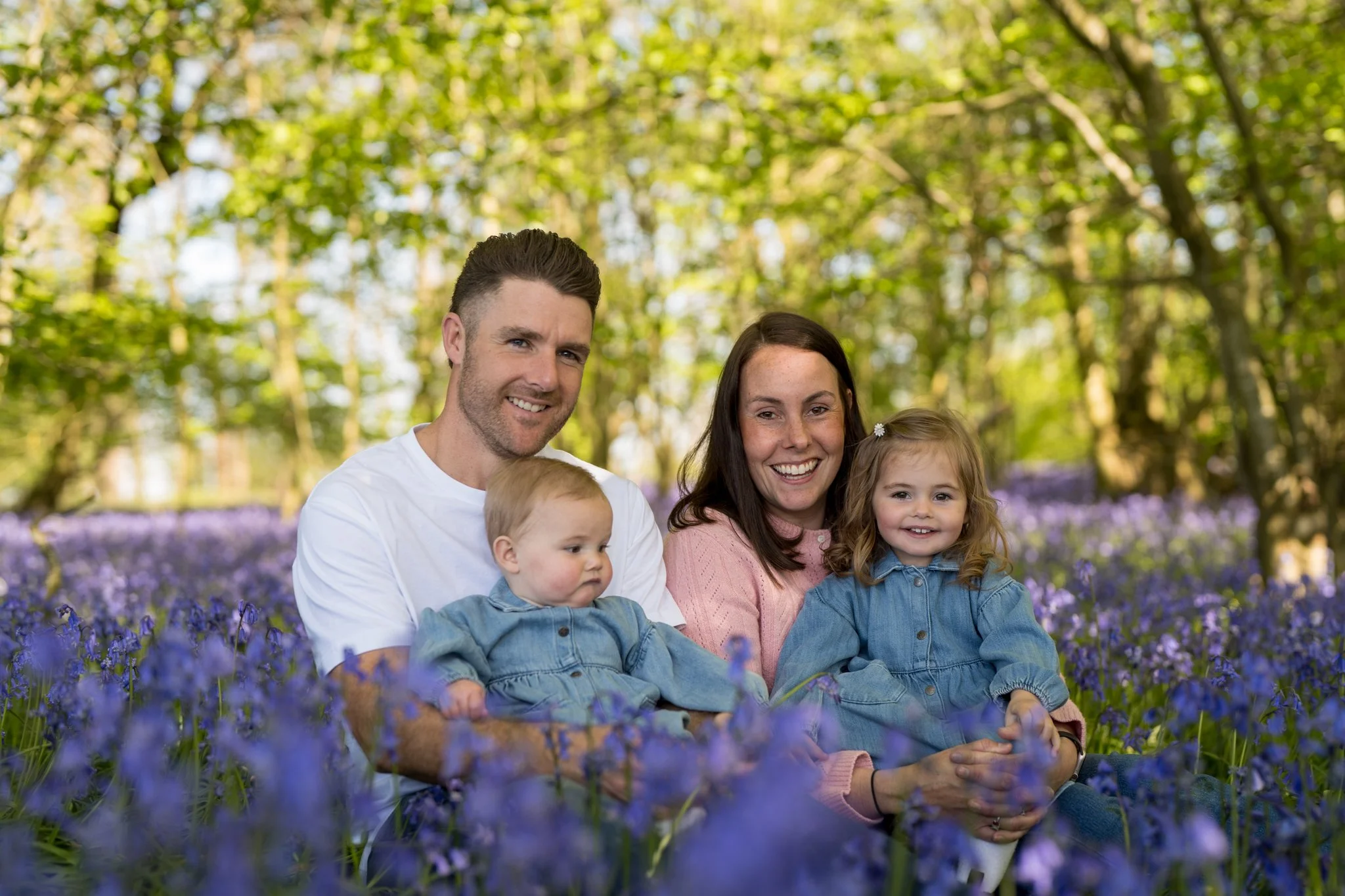 A family of four sitting outdoors in a forest of bluebell flowers, with green trees in the background, smiling and enjoying a sunny day.