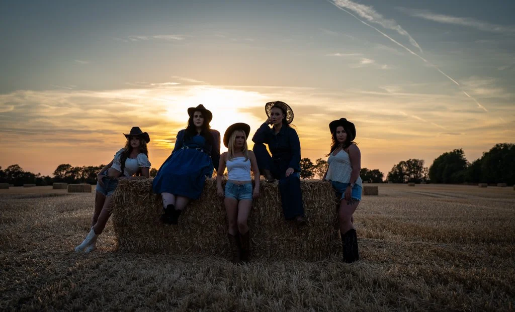 Five women in Western-style clothing and hats sitting and standing on a hay bale in an open field at sunset.
