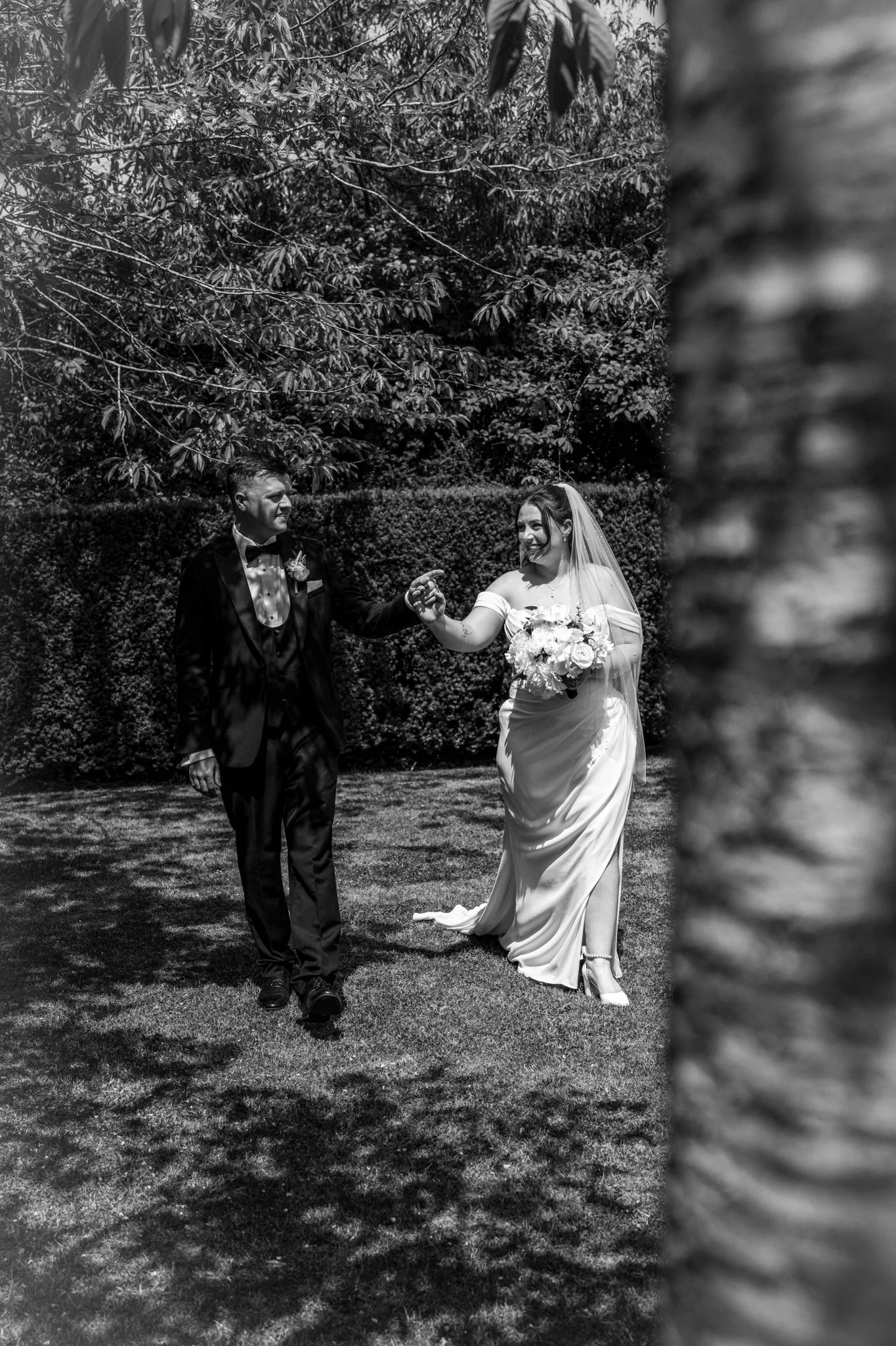 A black and white photo of a bride and groom holding hands in a garden. The groom is dressed in a tuxedo with a bow tie, and the bride is wearing a white wedding gown and veil, holding a bouquet of flowers. They are smiling at each other, with trees 