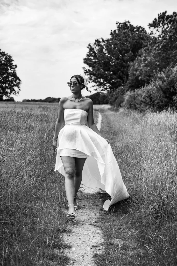 A woman in a strapless white dress walking on a dirt path through a grassy field with trees in the background, wearing sunglasses and sandals. Anne Wills Photography. Braintree town hall, Essex