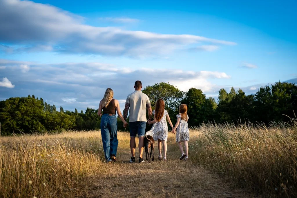 Family of five walking on a dirt path through a grassy field with trees and a partly cloudy sky in the background, holding hands.