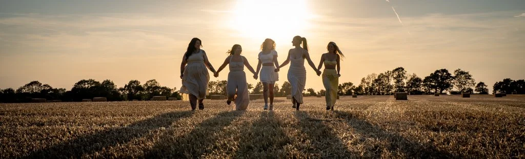 Six women holding hands and walking across a field during sunset.