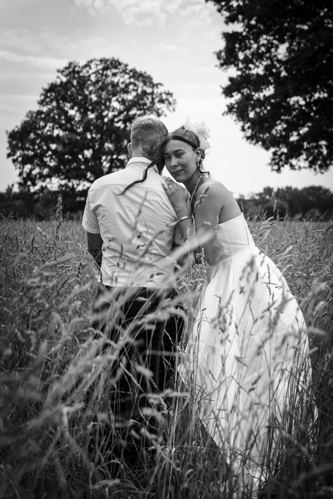 A black and white photo of a couple embracing in a field of tall grass with trees in the background. Anne Wills Photography. Braintree town hall, Essex