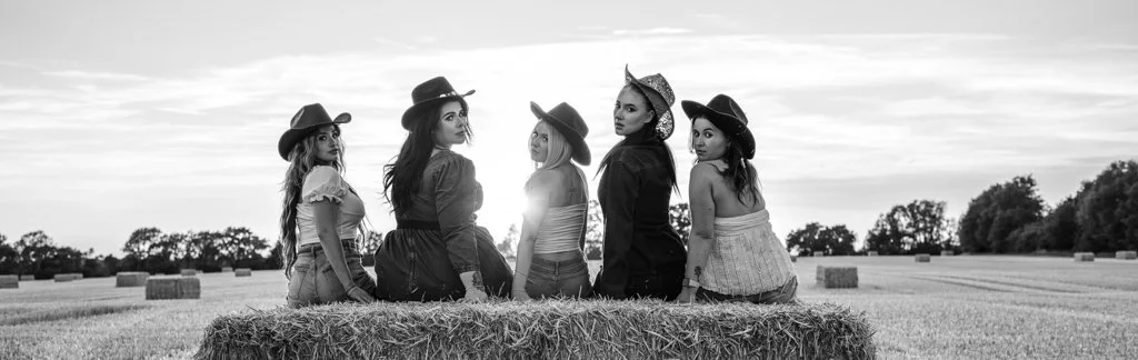 Five women wearing hats and casual outfits sitting on a hay bale in a field with trees in the distance, black and white photo.
