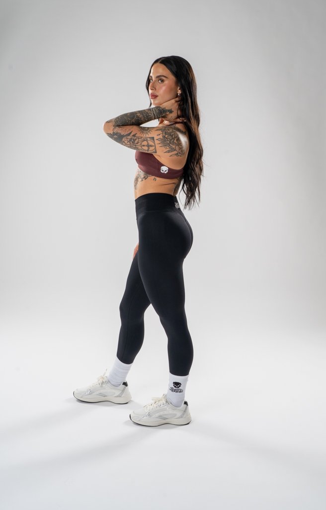 A fit woman with long dark hair and tattoos on her arms and side, wearing a maroon sports bra, black leggings, white socks, and white sneakers, standing against a plain white background. She is posing sideways with one hand on her neck.