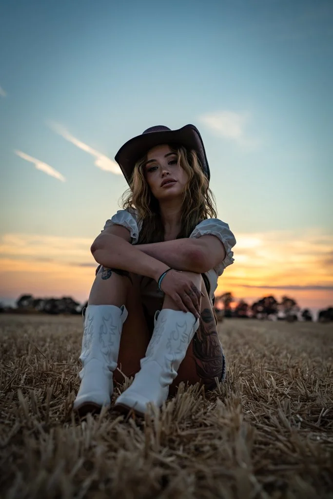 A young woman sitting in a field during sunset, wearing a wide-brimmed hat, white cowboy boots with writing, and a casual outfit, looking at the camera with a relaxed expression.