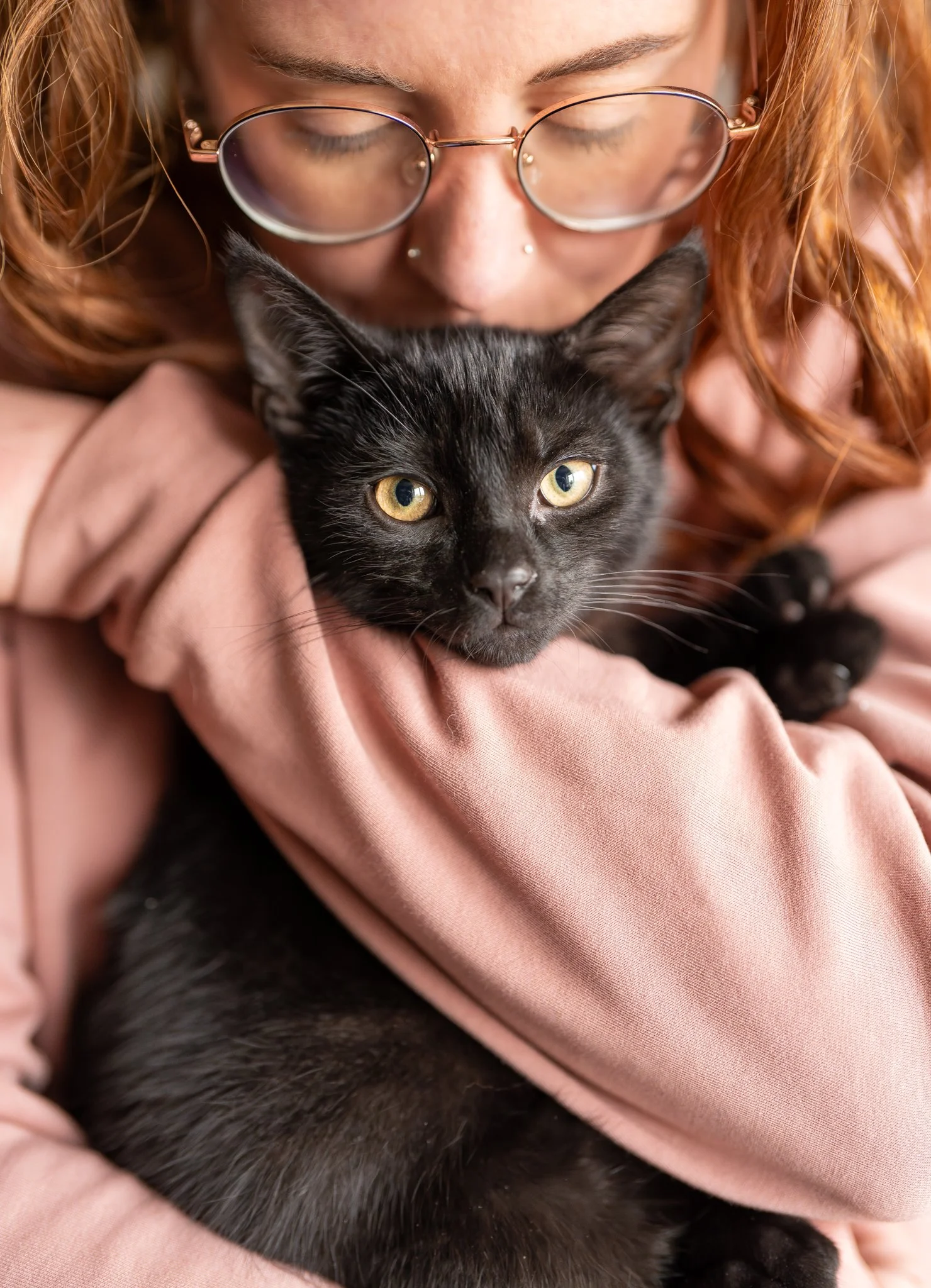 A woman with red hair and glasses holding a black kitten with yellow eyes close to her face. photographed in Rayne, essex. Anne Wills Photography 