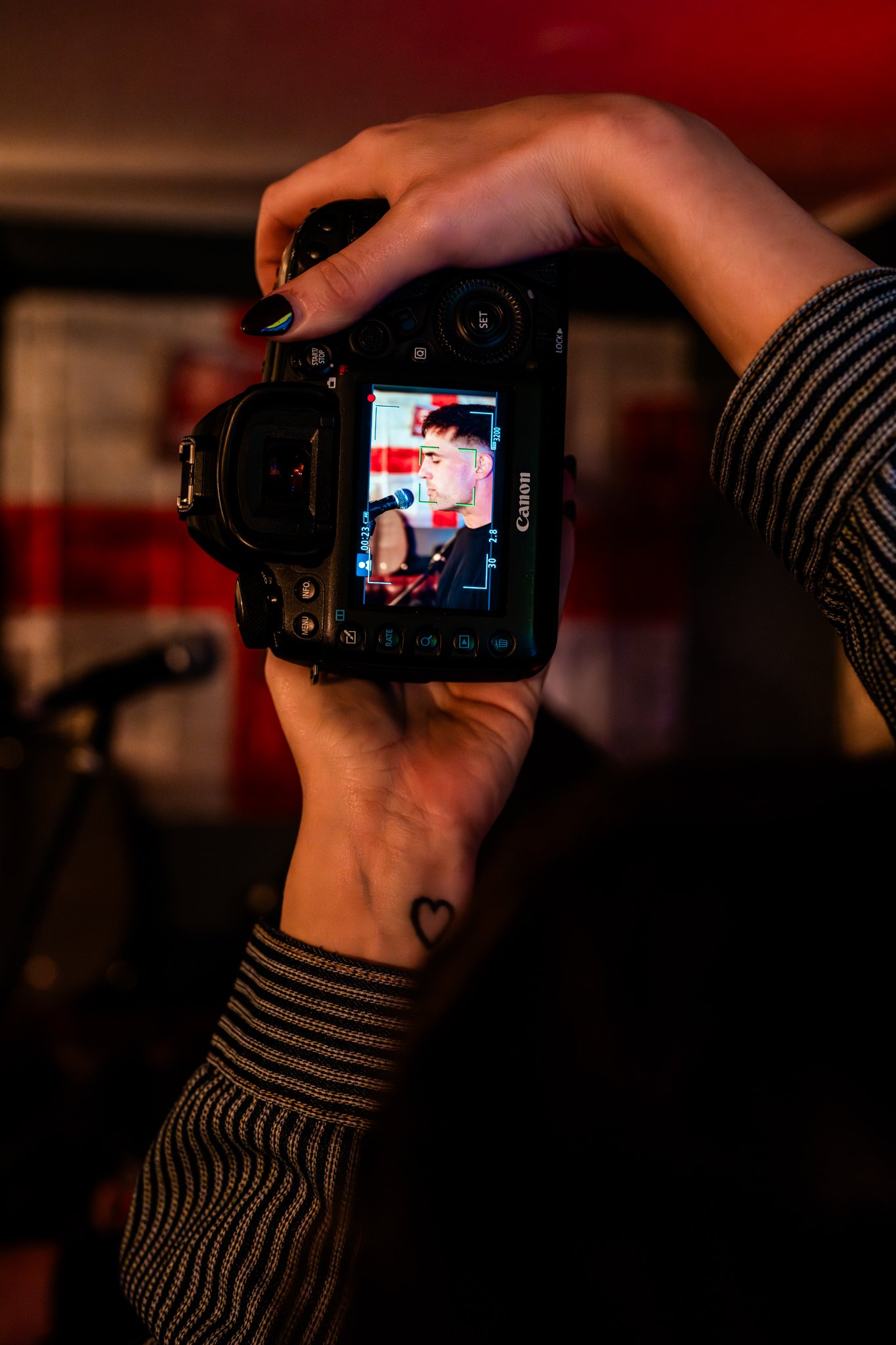 Person holding a Canon digital camera, capturing a woman speaking into a microphone, with a dark background and a tattoo of a heart on their wrist.