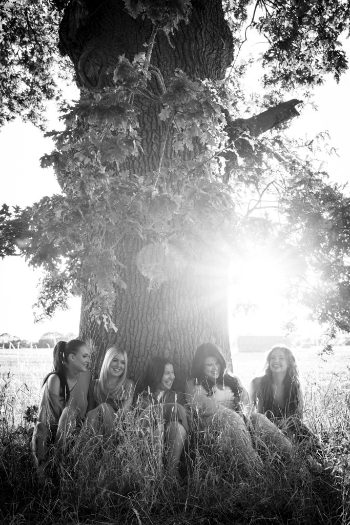 Five women sitting on grass under a large tree, smiling and talking.