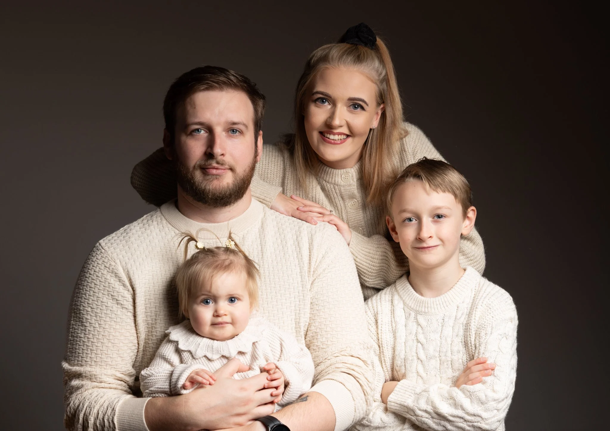 A family portrait with a man, woman, and three children, all wearing cream-colored knit sweaters, sitting close together against a dark background. Anne Wills Photography 