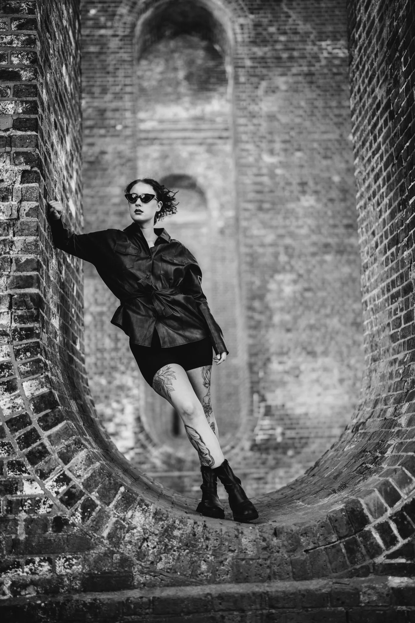 A woman with tattoos on her legs, wearing sunglasses, a black leather jacket, and black boots, posing against brick arches in a black and white photo. Photographed in colchester essex. Anne Wills Photography 