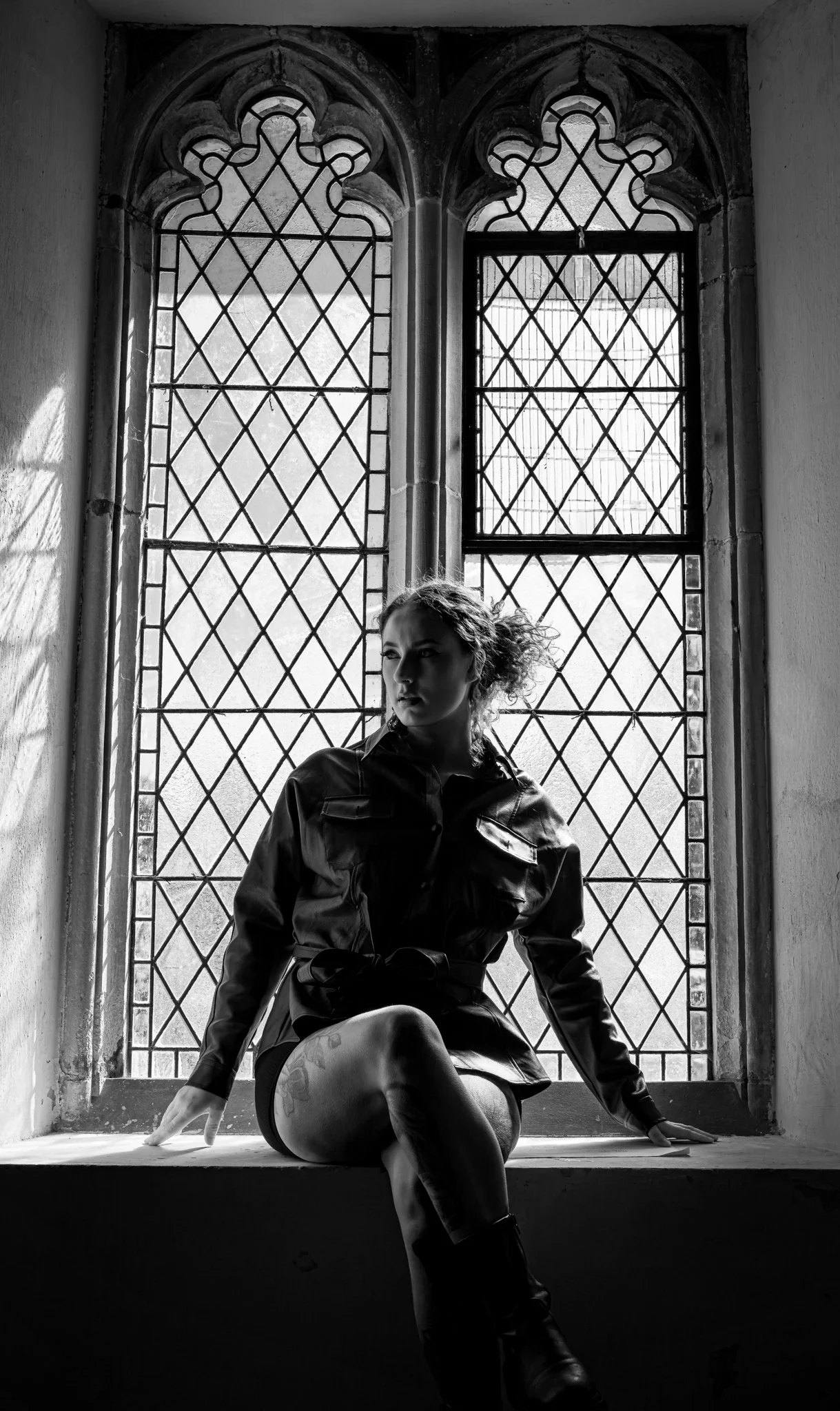 Black and white photo of a woman sitting on a windowsill inside a building with stained glass windows behind her, looking to the side with her hair partially masked by shadows.