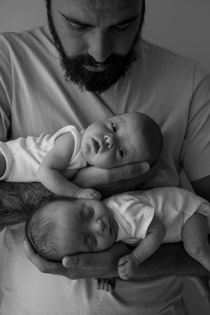 A bearded man holding two newborn babies, one awake looking at the camera, the other sleeping.