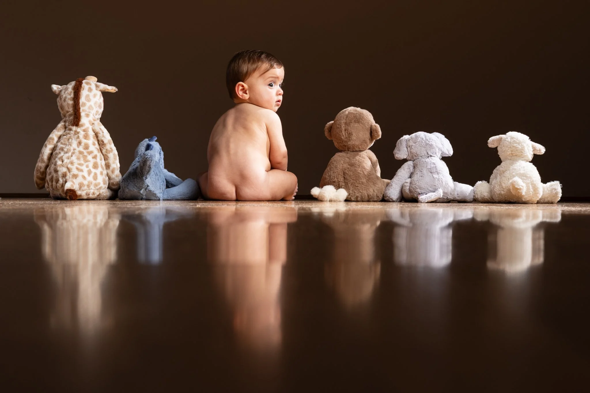 A baby sitting on a wooden floor surrounded by stuffed animal toys, with a dark brown background. Anne Wills Photography 
