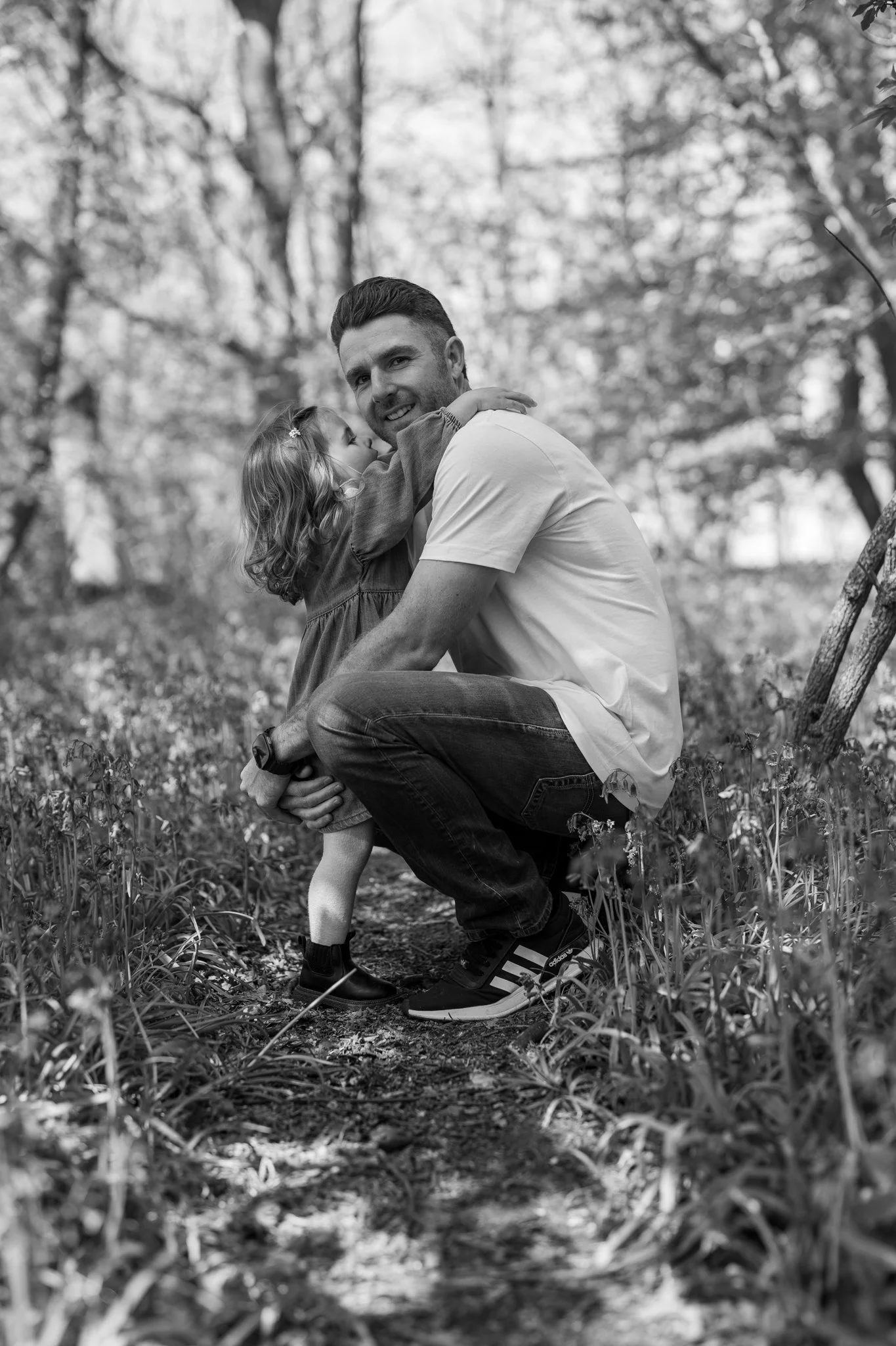 A man and young girl outdoors in a wooded area, smiling and hugging. The girl is kissing the man's cheek, and they are both kneeling close to the ground among grass and small plants. The background shows trees and leaves. Bluebell woods, Salford, Ess