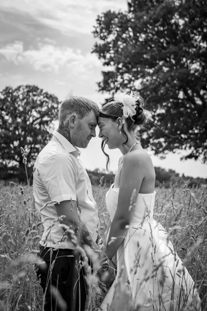A black and white photo of a couple standing close together in a grassy field, with their foreheads touching and smiling. The woman is wearing a strapless dress and a floral hair accessory, and the man is in a short-sleeved shirt. trees are visible i