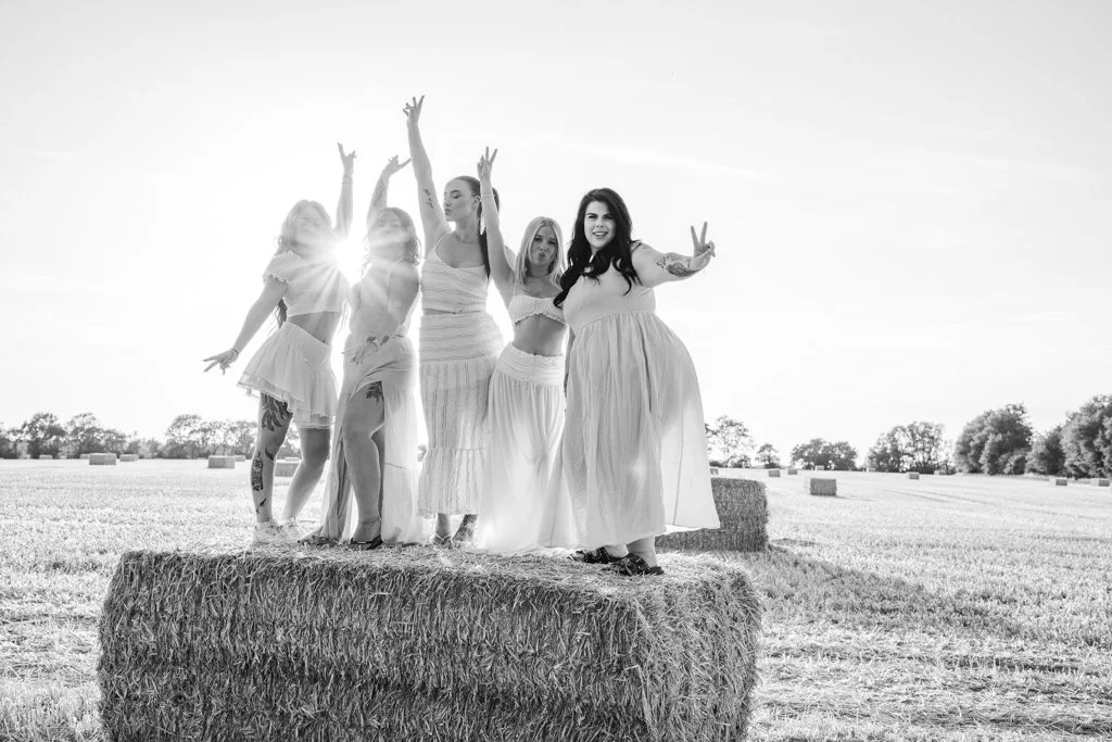 Five women standing on a hay bale in a farm field, smiling and making peace signs, dressed in light-colored dresses on a sunny day.