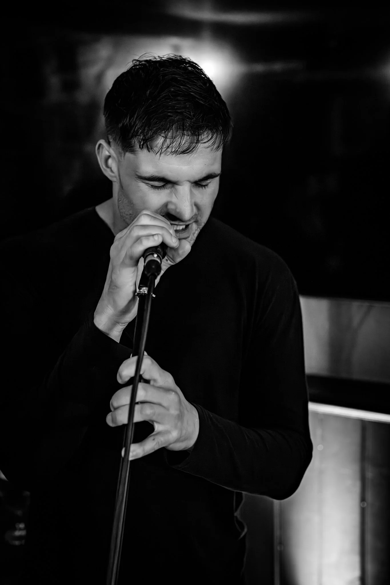 A young man with dark, wet hair singing into a microphone with his eyes closed, in a dimly lit setting.