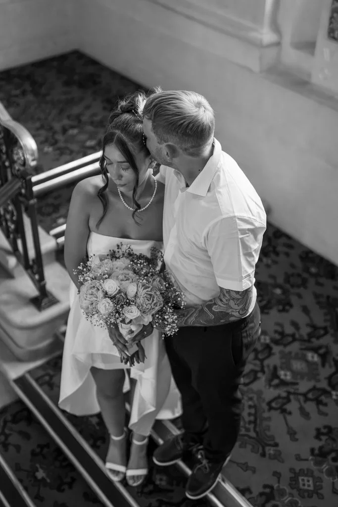 A couple on their wedding day standing on a staircase, with the bride holding a bouquet of flowers and the groom kissing her forehead. Anne Wills Photography. Braintree town hall, Essex