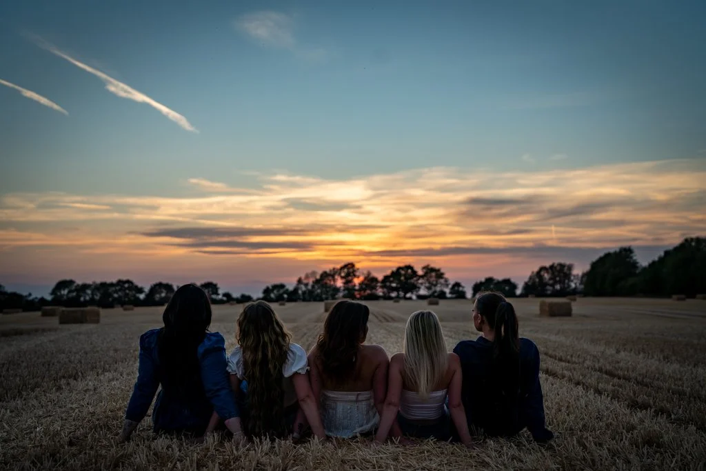 Six women are sitting on a hay field, watching a sunset with scattered clouds and trees in the background.