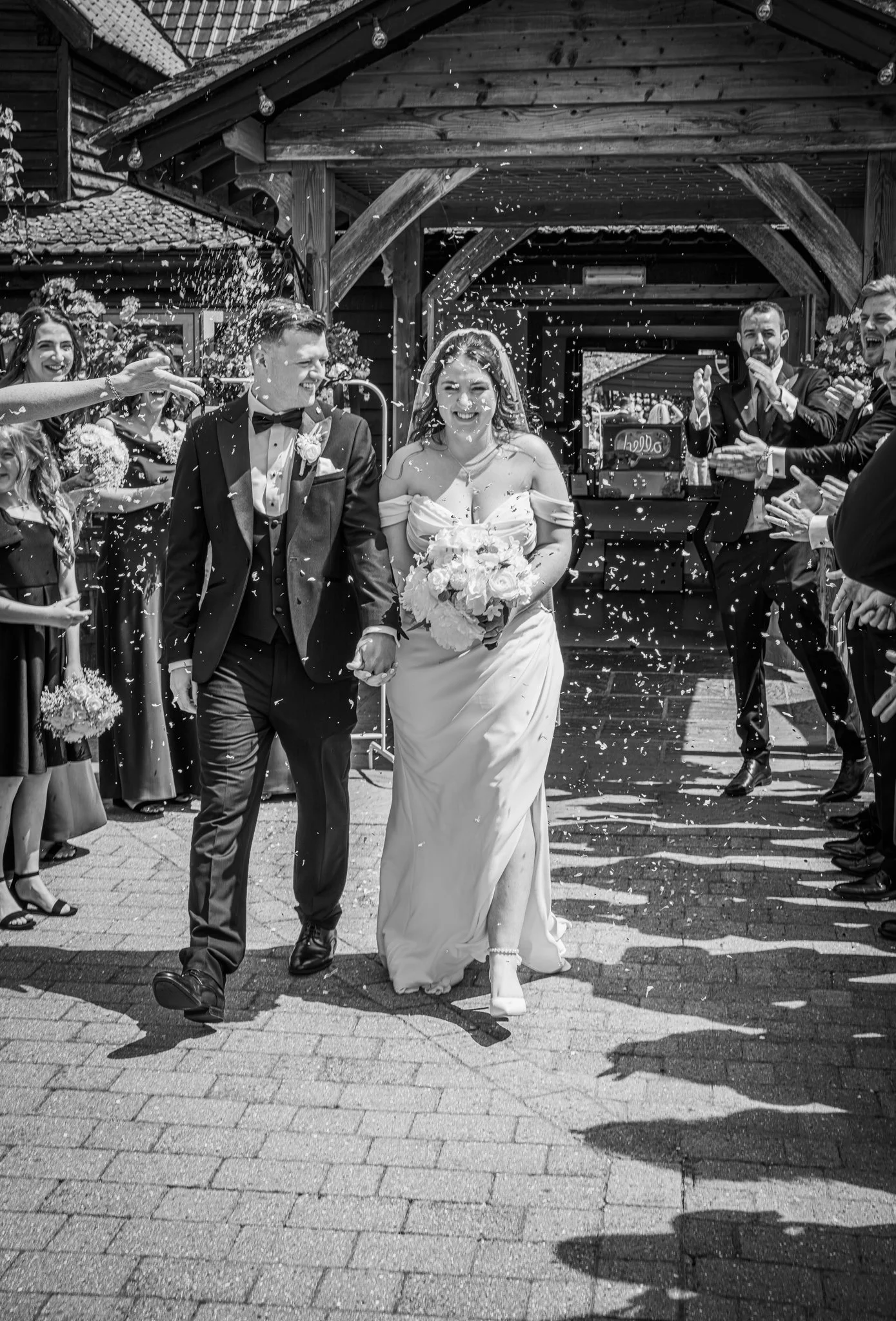 A bride and groom walk hand in hand outdoors, smiling as guests throw confetti in celebration, with some guests clapping and cheering.