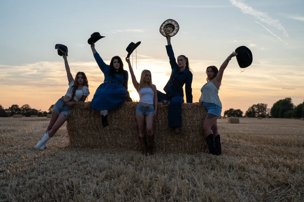 Group of five young women in casual summer clothes and cowboy hats posing next to a hay bale in a field at sunset.