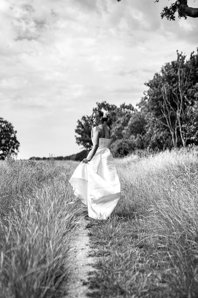 A woman in a strapless dress holding its hem, walking on a narrow dirt path through tall grass in a natural outdoor setting with trees and cloudy sky. Anne Wills Photography. Braintree town hall, Essex