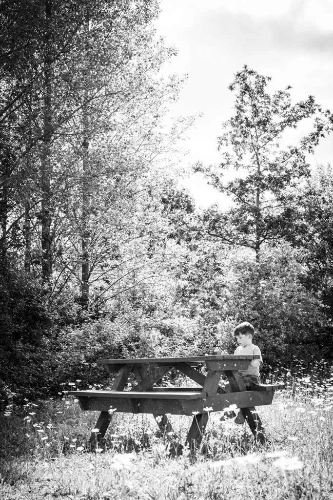 A young boy sitting alone at a wooden picnic table in a grassy field surrounded by trees, with sunlight filtering through the foliage. Anne Wills Photography. Braintree, Essex