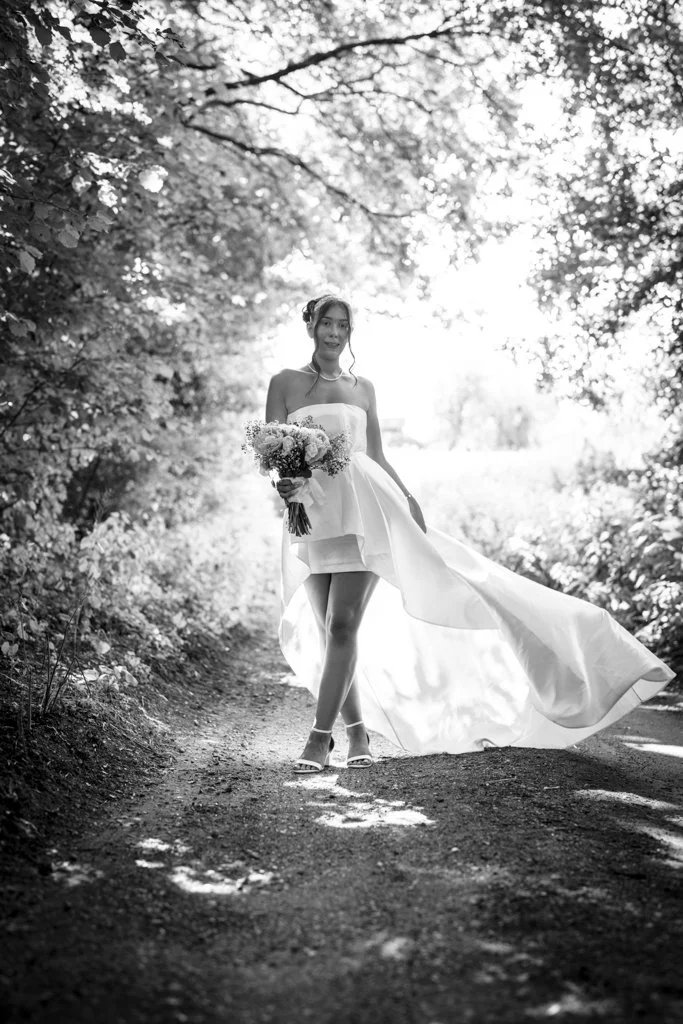 A bride in a strapless wedding gown walking on a wooded forest path, holding a bouquet of flowers and lifting her dress slightly, sunlight filtering through the trees. Anne Wills Photography. stabbing, essex