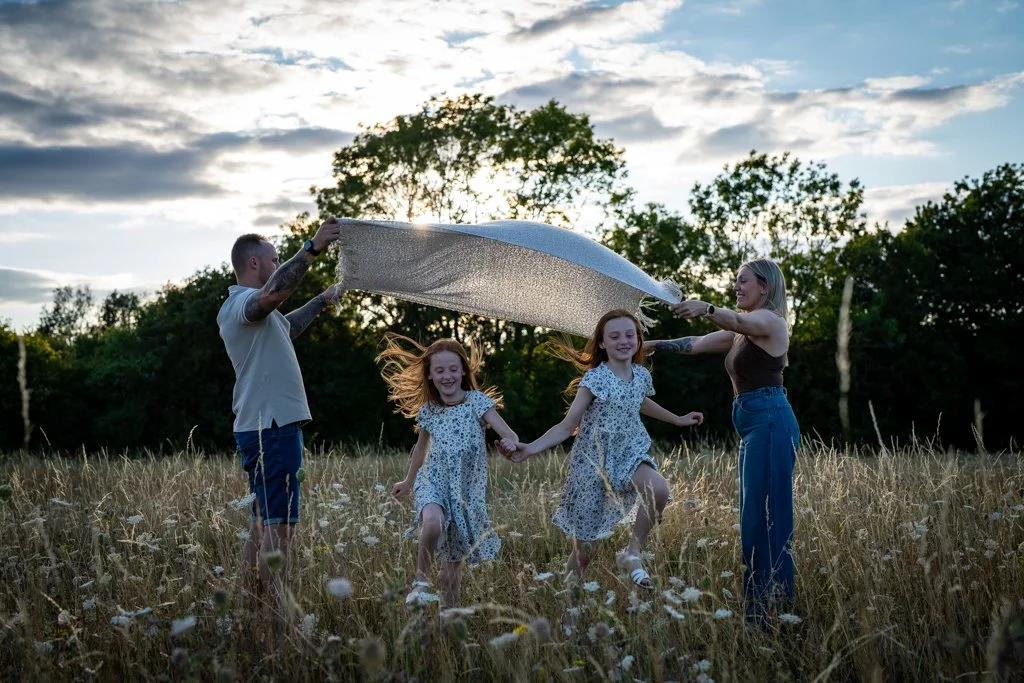 Family of four holding a blanket over their children in a field with trees in the background at sunset.