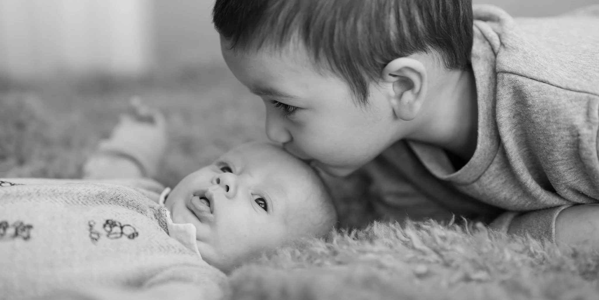 A young boy leaning over and giving a gentle kiss to a baby lying on a fuzzy blanket. Photographed in customers home in Chelmsford, Essex. Anne Wills Photography 