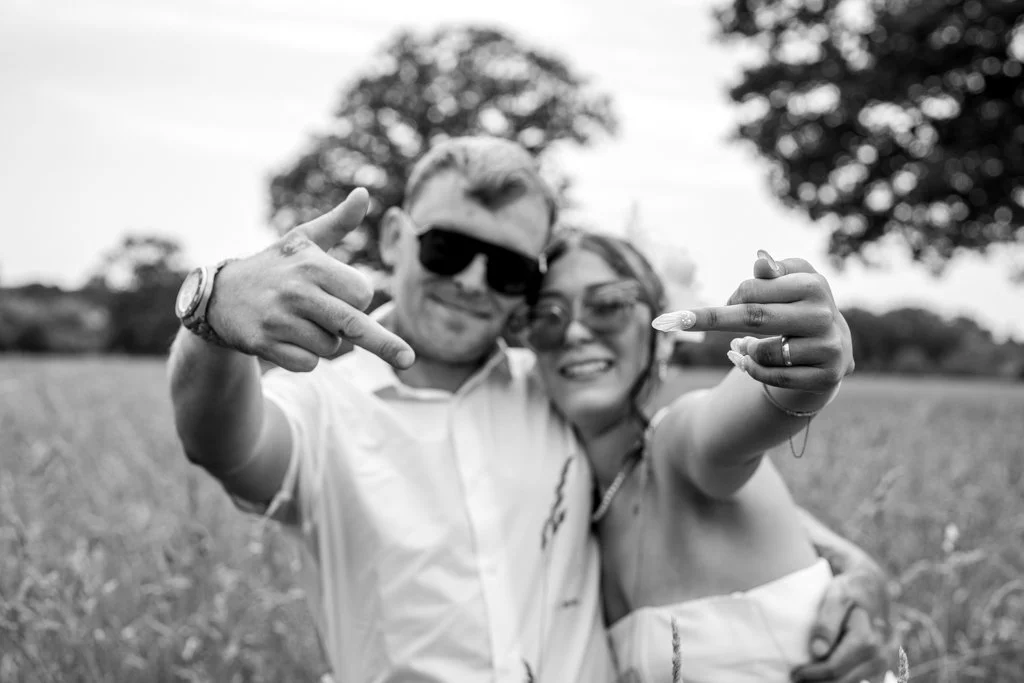 Pair of people smiling and making hand gestures in an outdoor field, with trees in the background, black and white photo. Anne Wills Photography. Braintree, essex