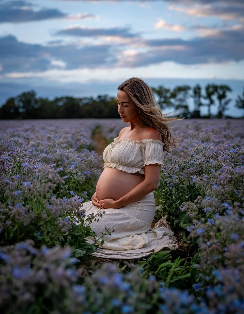 Pregnant woman in a white dress sitting in a field of purple flowers during sunset.