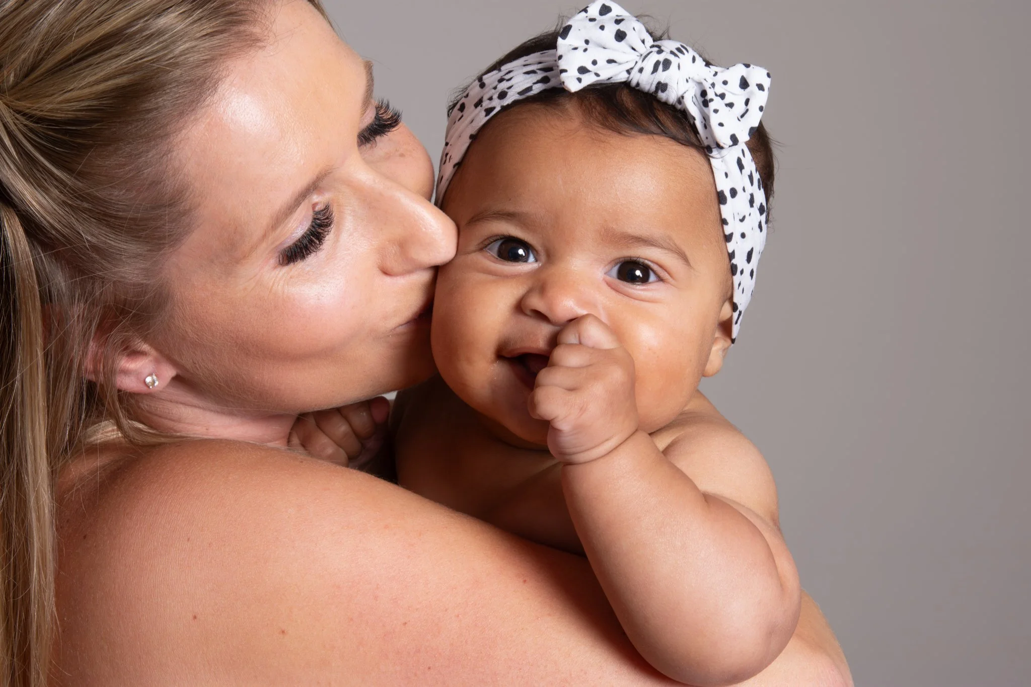 A woman with long blonde hair, earrings, and makeup holds a smiling baby girl with dark hair and big brown eyes, wearing a white headband with black polka dots, against a plain gray background. Anne Wills Photography 
