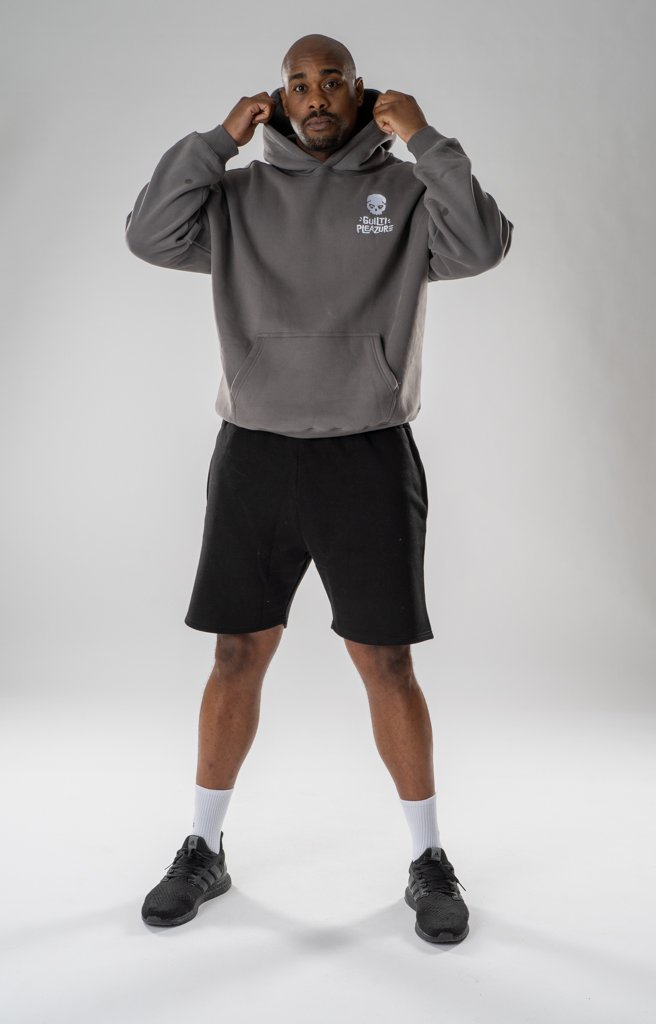 Man adjusting the collar of his gray hoodie, wearing black shorts, white socks, and black athletic shoes, standing against a plain white background.