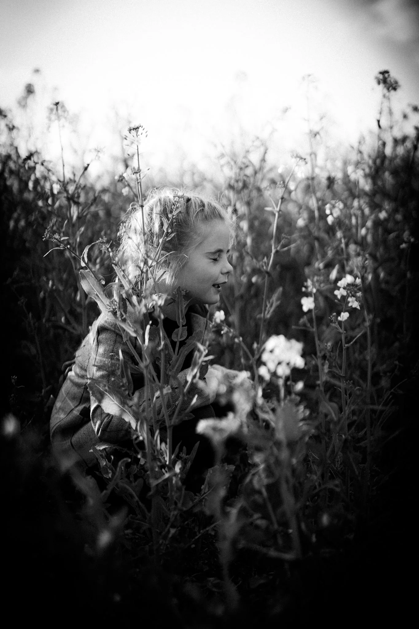 A young girl with closed eyes and a smile is sitting among tall plants and flowers in a field, with sunlight shining brightly overhead in a black and white photograph. photographed in felsted essex. Anne Wills Photography 