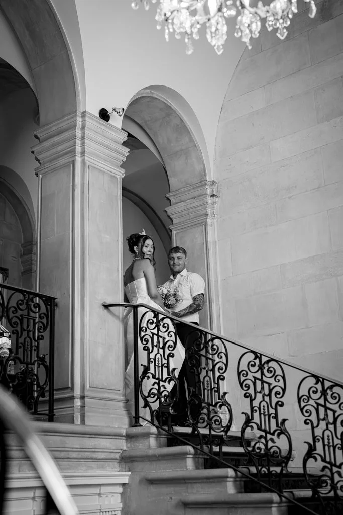 A couple standing on a staircase with ornate railing inside a grand building, possibly a church or mansion, with high ceilings, arches, and a chandelier, in black and white. Anne Wills Photography. Braintree town hall, Essex