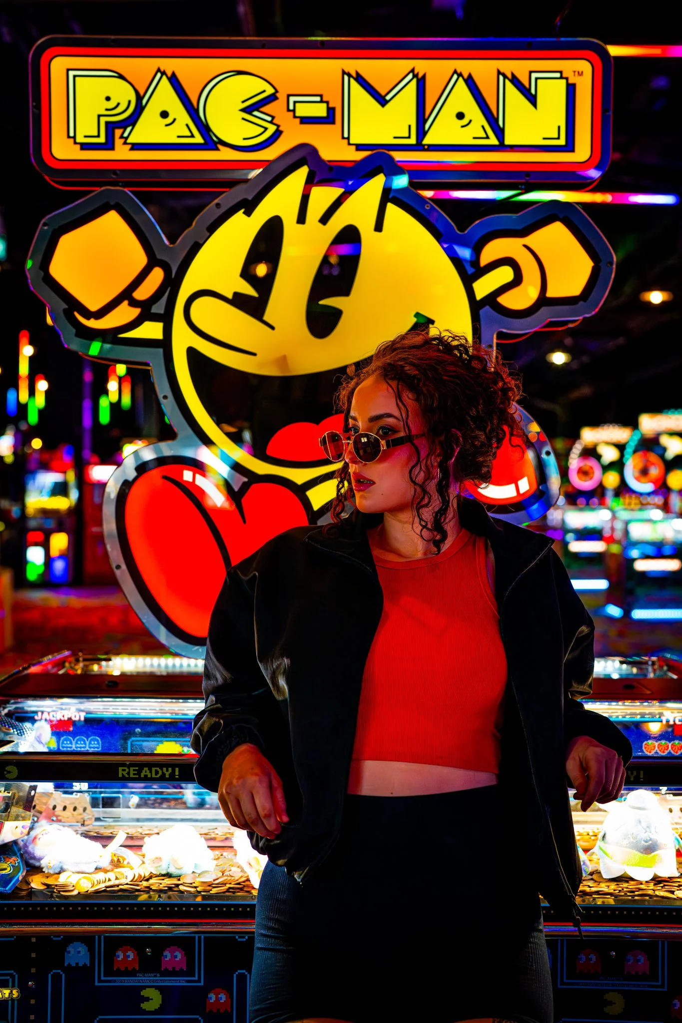 A woman with curly hair wearing sunglasses and a black jacket stands in front of a neon Pac-Man sign at a colorful arcade or amusement park during nighttime. photographed in Walton, essex in the arcades on the pier. Anne Wills Photography 