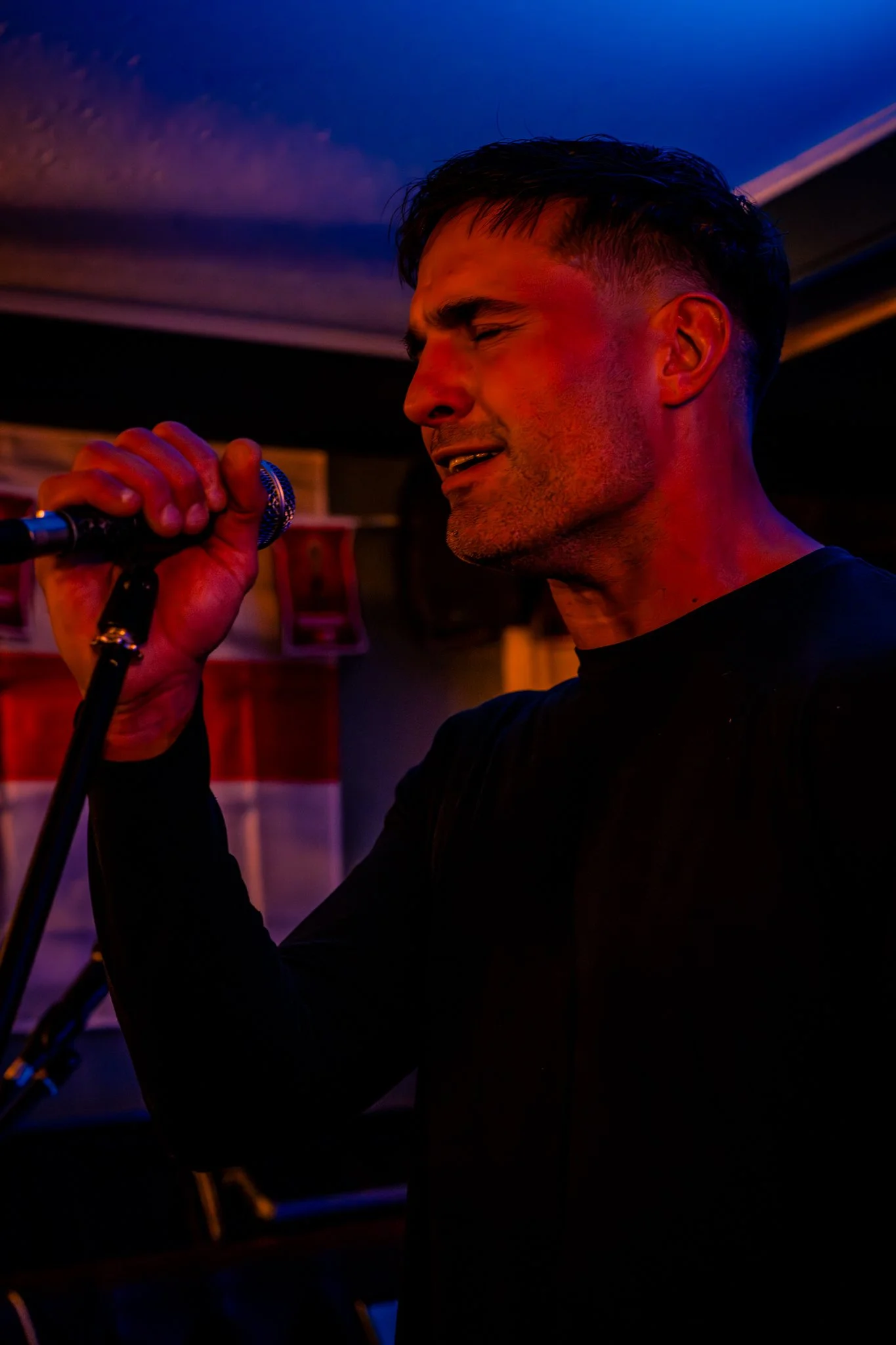A man singing into a microphone with a dark background, illuminated by colored stage lights. Anne Wills Photography. Braintree, Essex