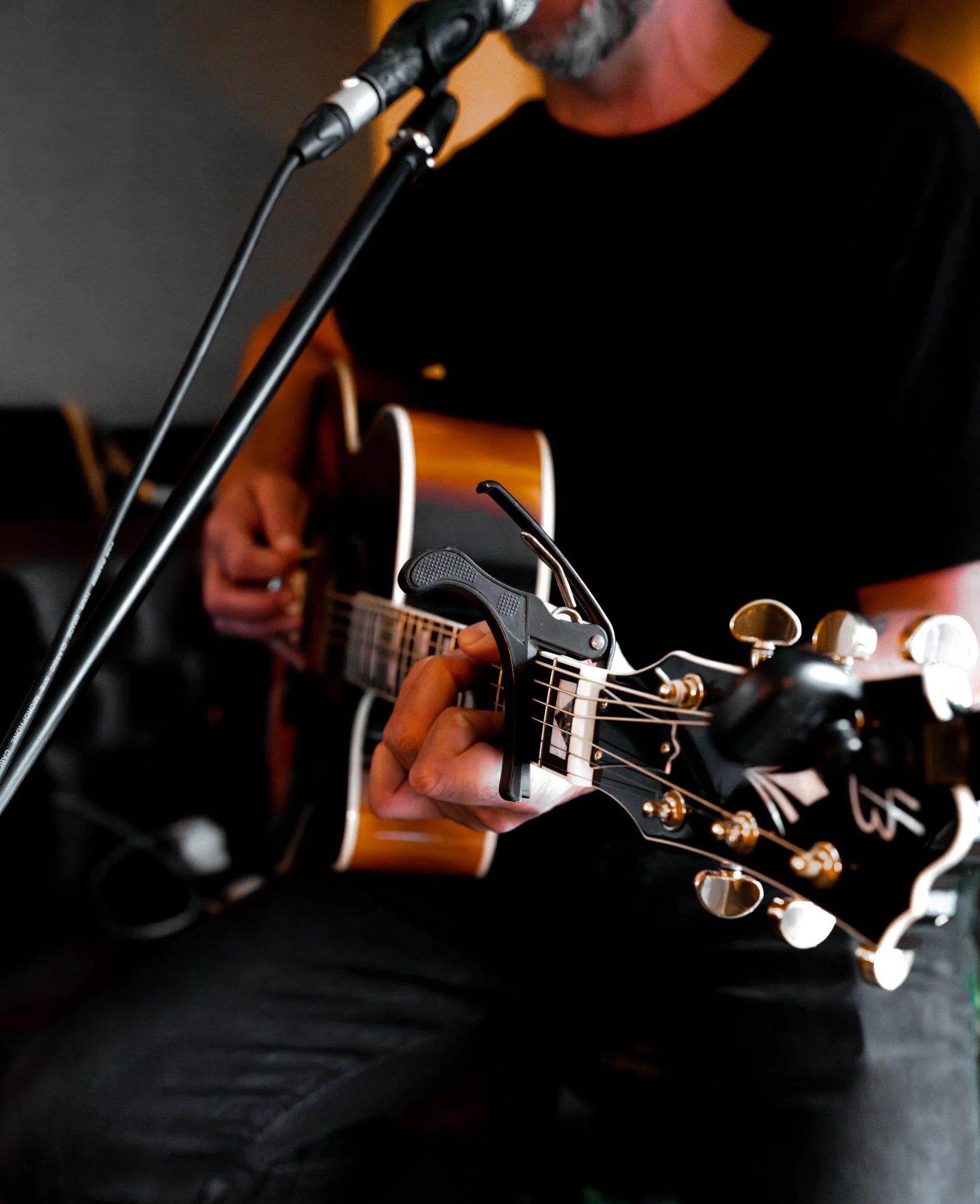 Close-up of a person playing an acoustic guitar, with focus on their hand on the fretboard and part of their face, microphone and guitar keys visible, dark background.
