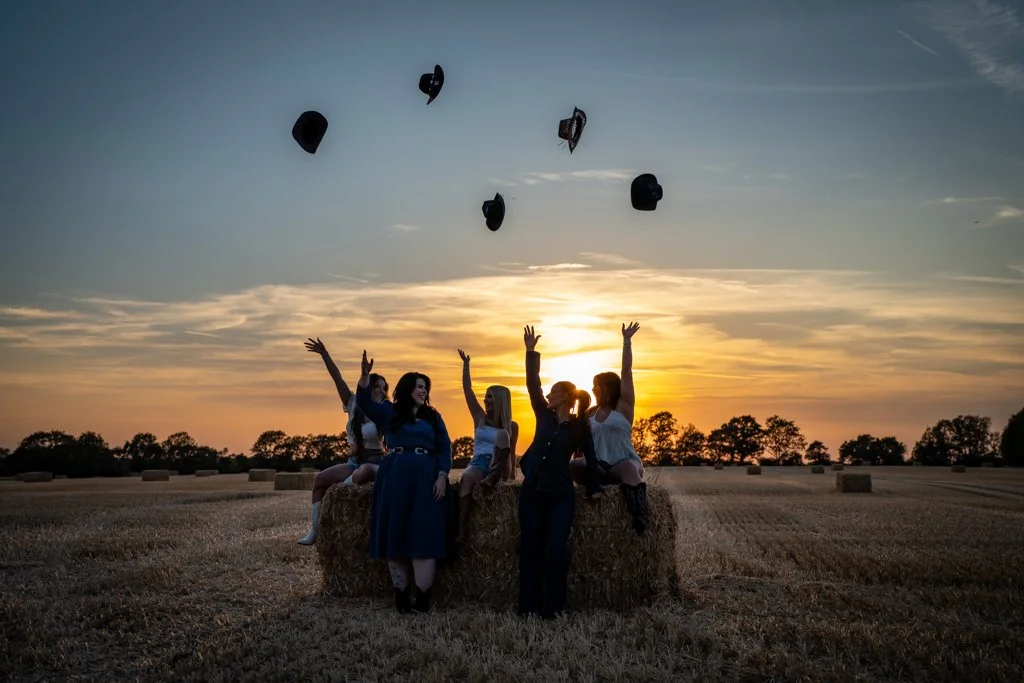 Group of women celebrating on a hay bale during sunset in a field, tossing their hats into the air.