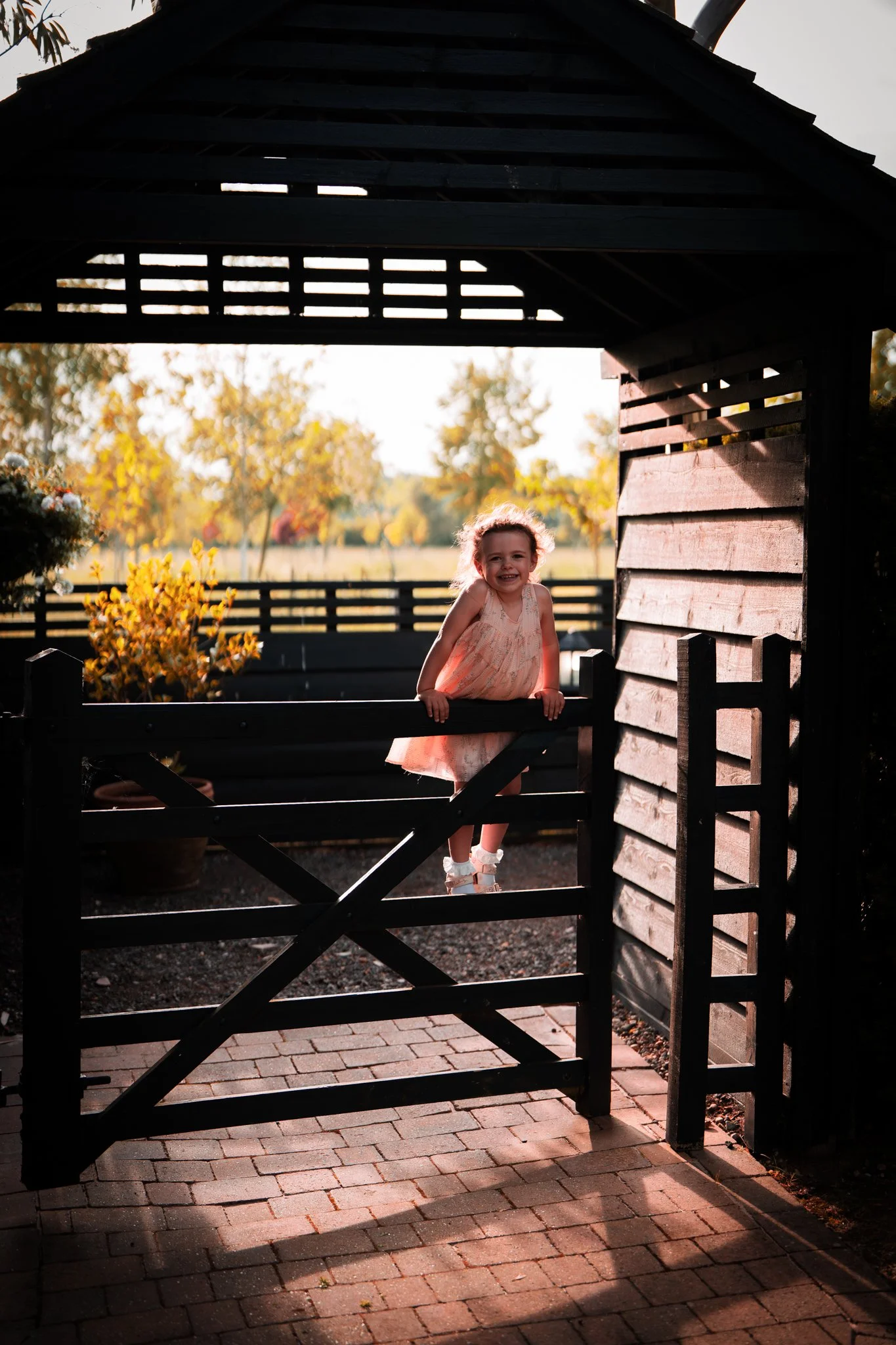 A young girl with curly hair smiling and leaning on a wooden gate in a garden, with autumn trees and sunlight in the background. Anne Wills Photography. Maidens Barn wedding venue, Essex