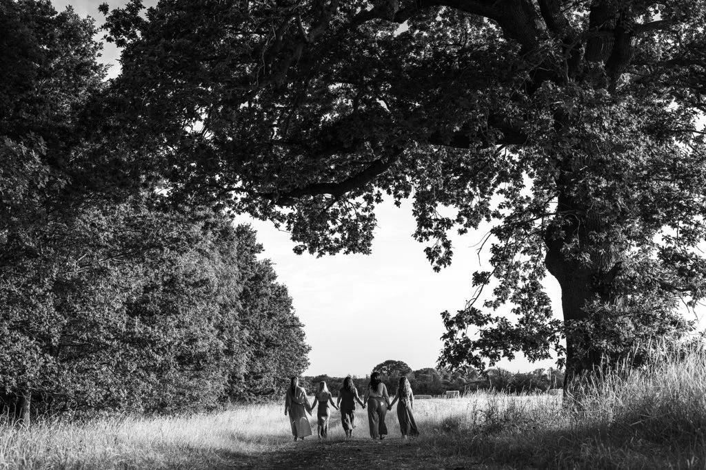 Four women holding hands walk along a trail through a park with large trees.