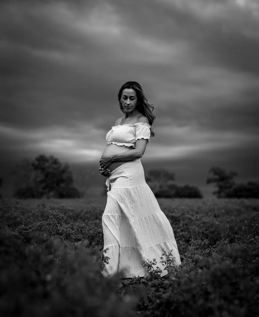 A pregnant woman stands in a field with trees in the background under a cloudy sky, wearing a flowing off-the-shoulder dress, smiling softly, with her hands resting on her belly.