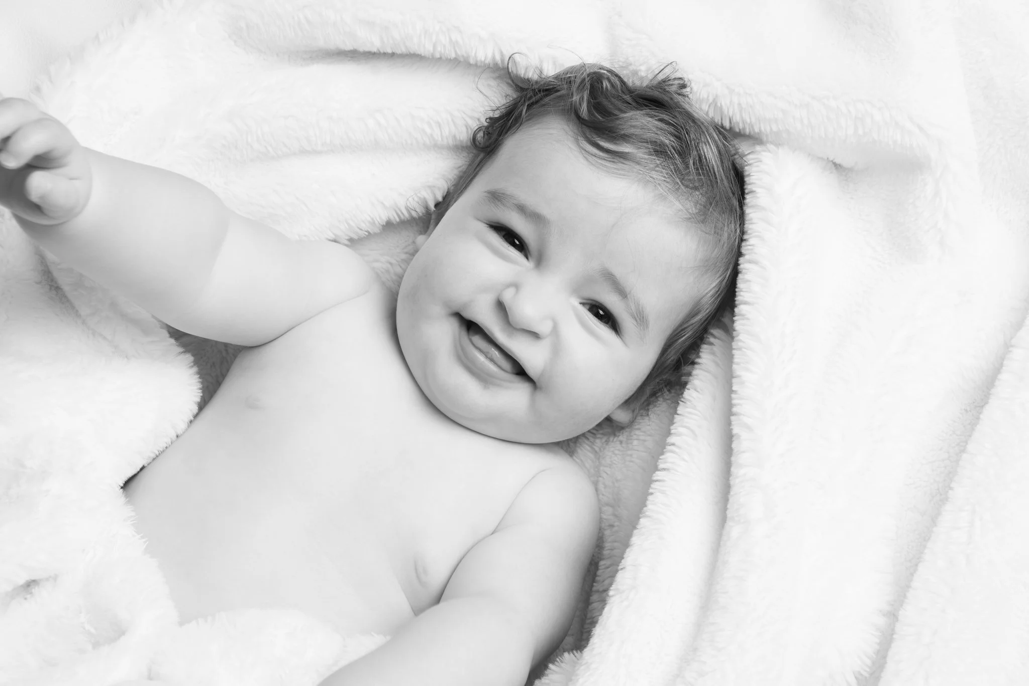 Black and white photo of a smiling toddler lying on a soft, fluffy blanket with one arm extended toward the camera. Photographed in a customers home in Braintree Essex. Anne Wills Photography 