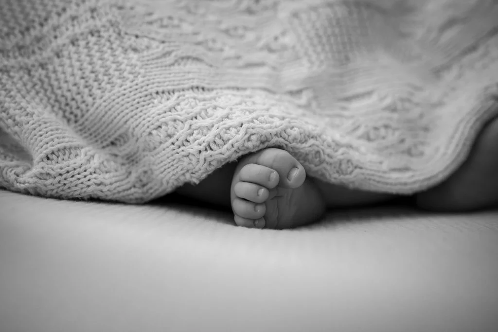 Close-up of a baby's hand gripping the edge of a knitted blanket, with the rest of the body hidden under the blanket.