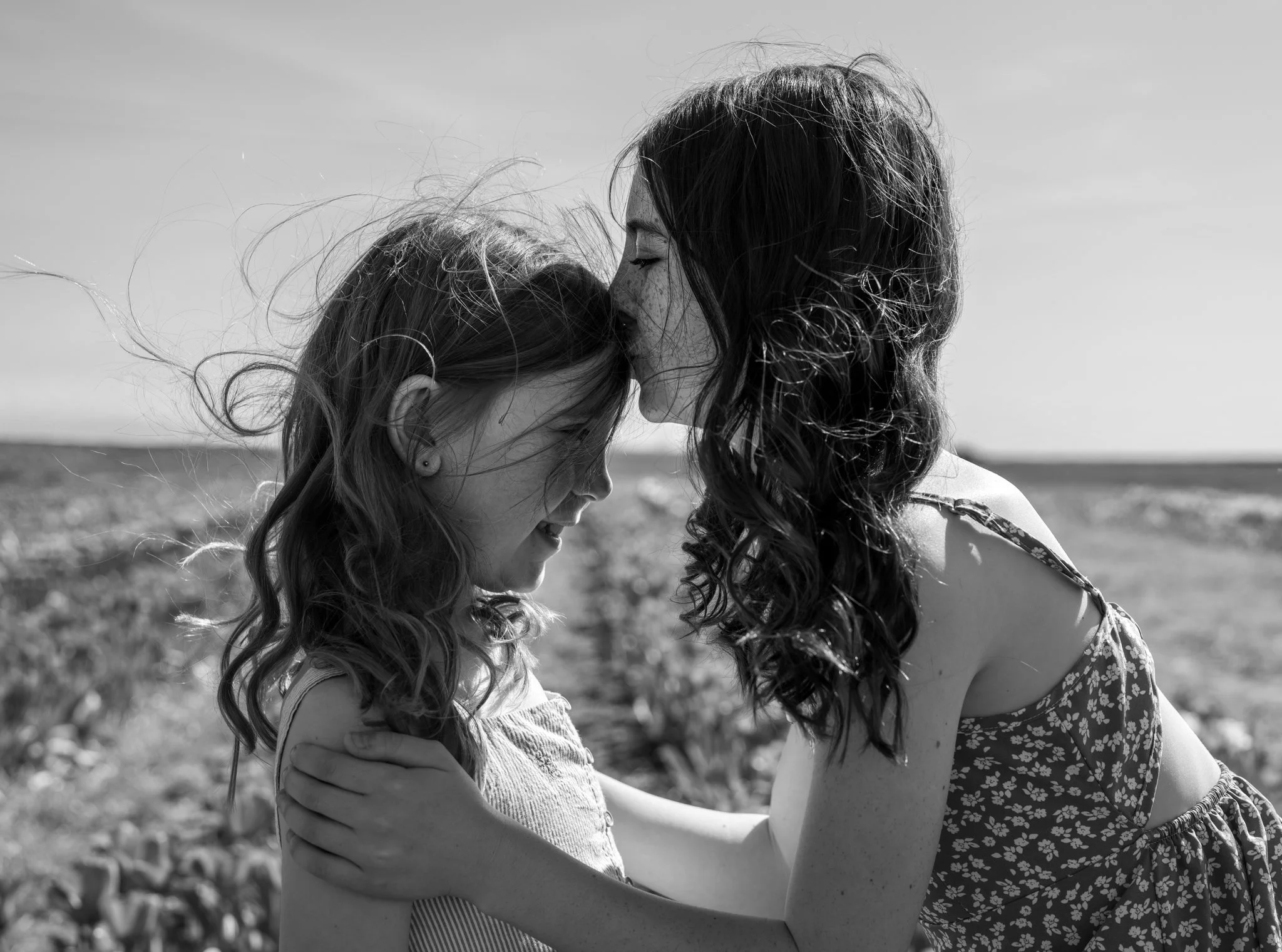 A woman and a girl with wavy hair are standing close together outdoors, facing each other, with their foreheads touching and eyes closed, sharing an intimate moment. The woman is wearing a floral dress, and the girl is in a sleeveless top. Their arms